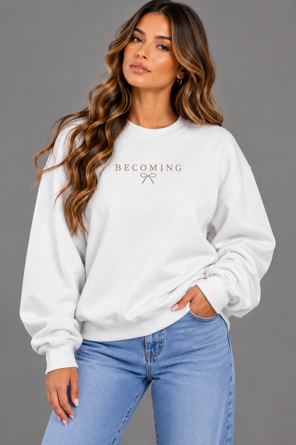 A woman wearing a white crewneck sweatshirt with 'BECOMING' and a small bow graphic printed in gold on the chest, paired with light blue jeans, against a solid gray studio background.