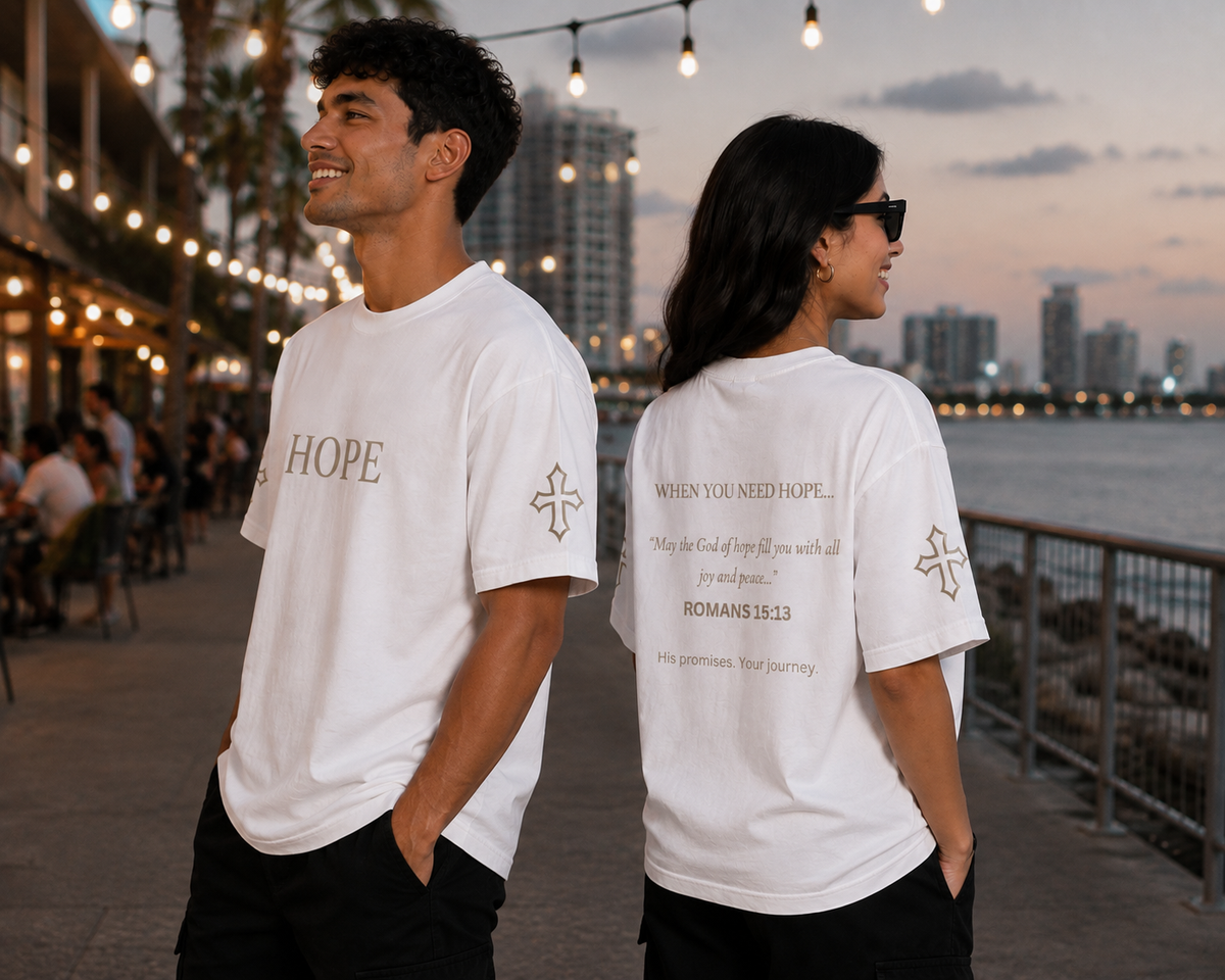 Male and female models wearing white Hope embroidered T-shirts from the Promise Collection, featuring embroidered chest and sleeve details with Romans 15:13 scripture printed on the back, photographed by the waterfront at sunset.