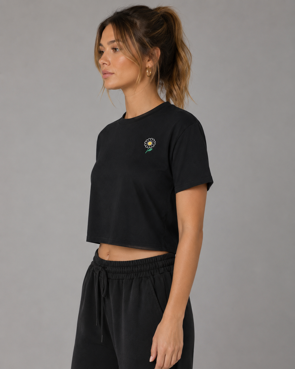 Side view of woman modeling a black cropped T-shirt with a small embroidered daisy on the chest, paired with black jogger pants.
