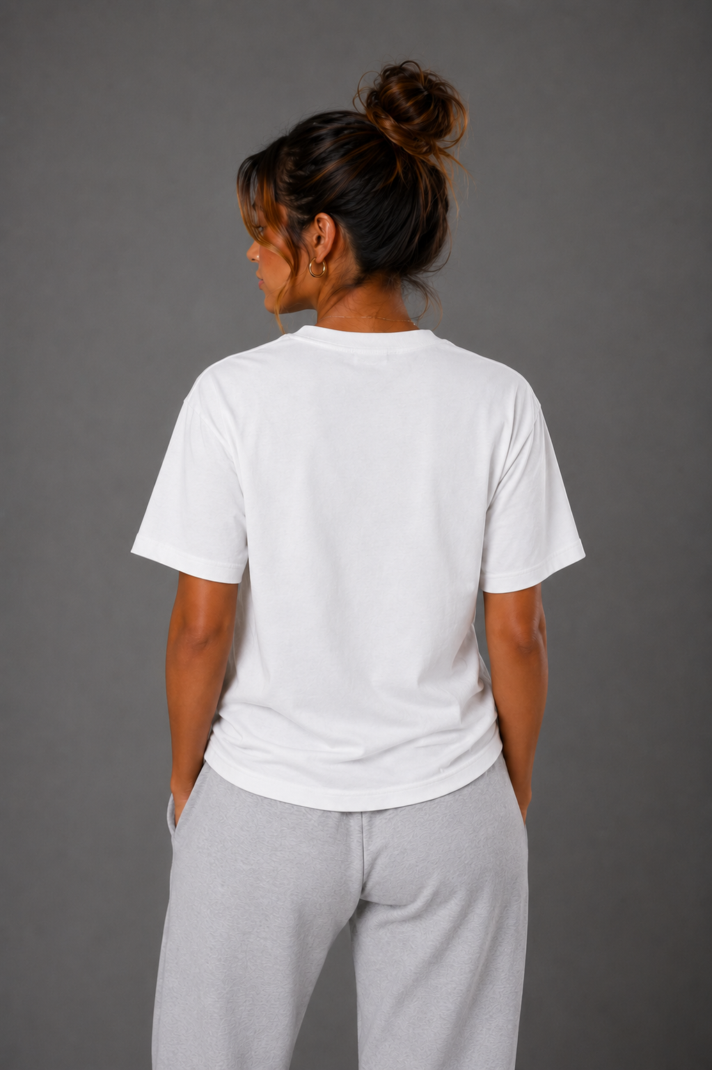 Woman modeling the back of a white T-shirt with gray sweatpants against a gray studio background.