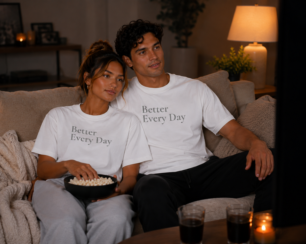 Couple relaxing at home in matching white “Better Every Day” motivational T-shirts while watching TV and sharing popcorn.