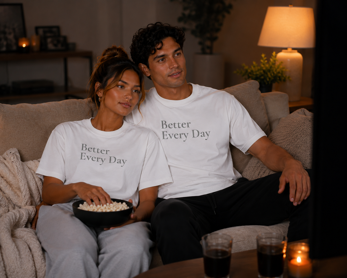 Couple relaxing at home in matching white “Better Every Day” motivational T-shirts while watching TV and sharing popcorn.