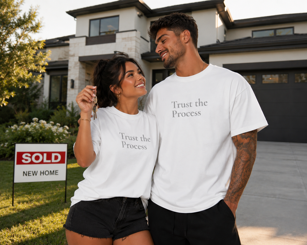 Happy couple wearing matching white “Trust the Process” motivational T-shirts celebrating in front of their newly purchased home.