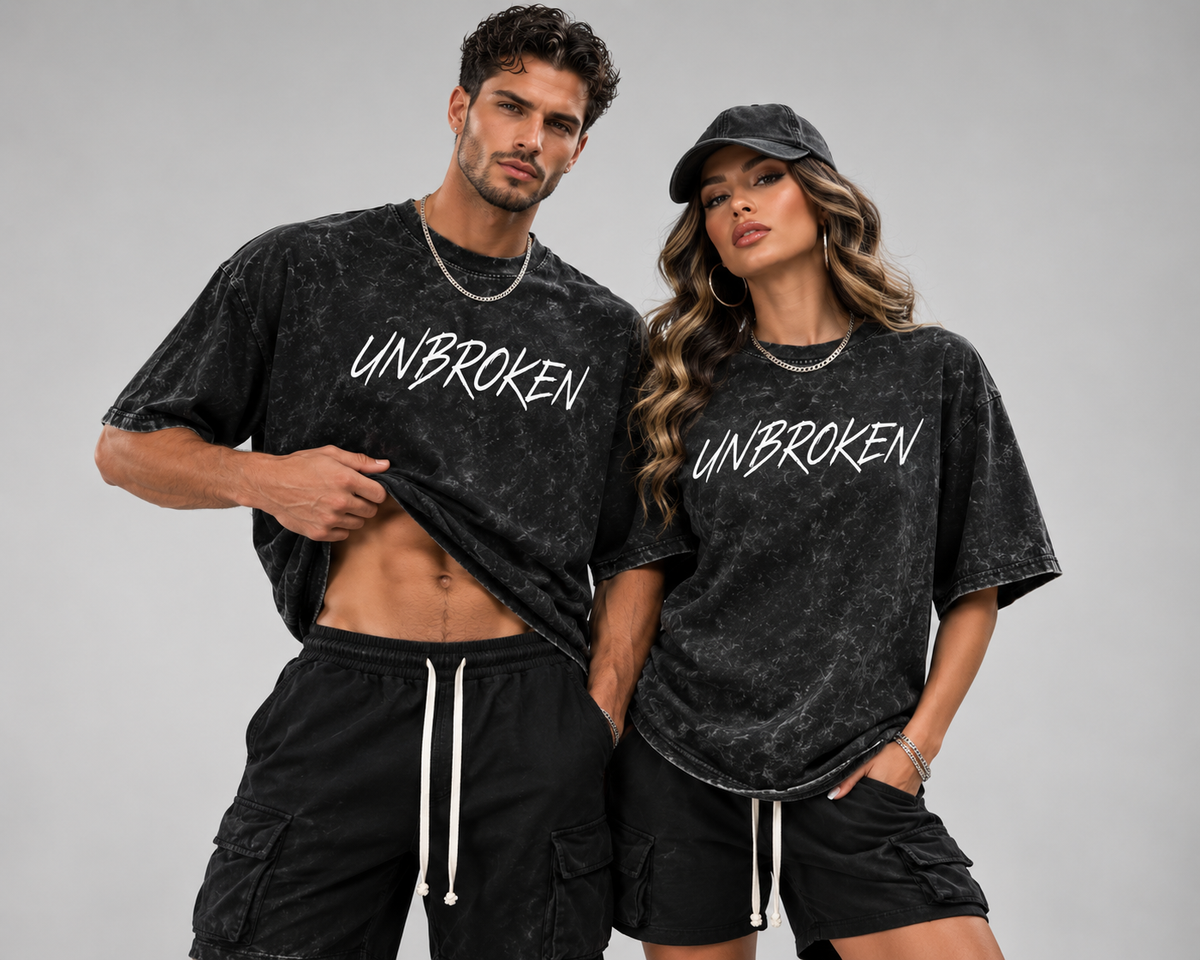 Man and woman modeling matching black acid-wash “UNBROKEN” graphic T-shirts with a bold streetwear look against a gray studio background.