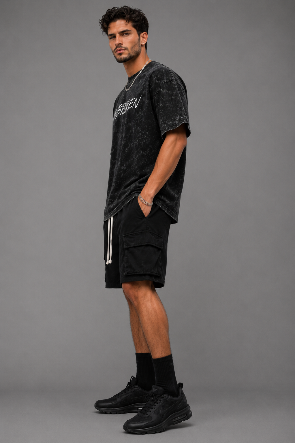 Man modeling a black acid-wash “UNBROKEN” oversized graphic T-shirt with black cargo shorts against a gray studio background.