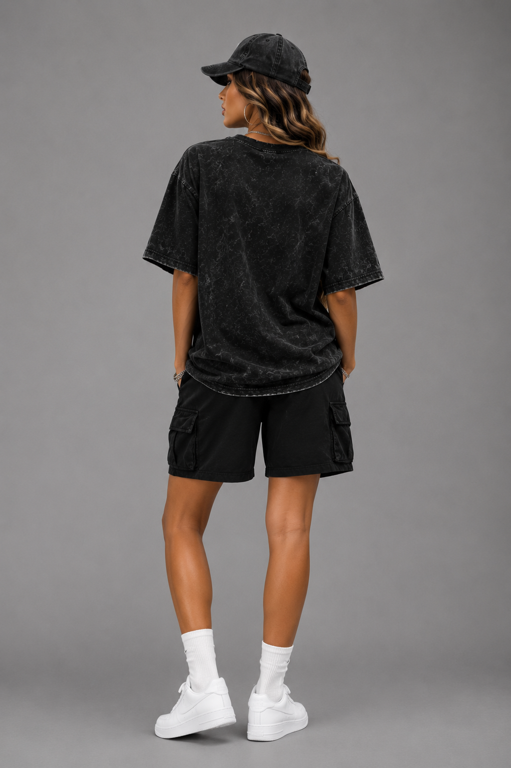 Woman modeling the back of a black acid-wash oversized T-shirt with black cargo shorts and a matching cap against a gray studio background.