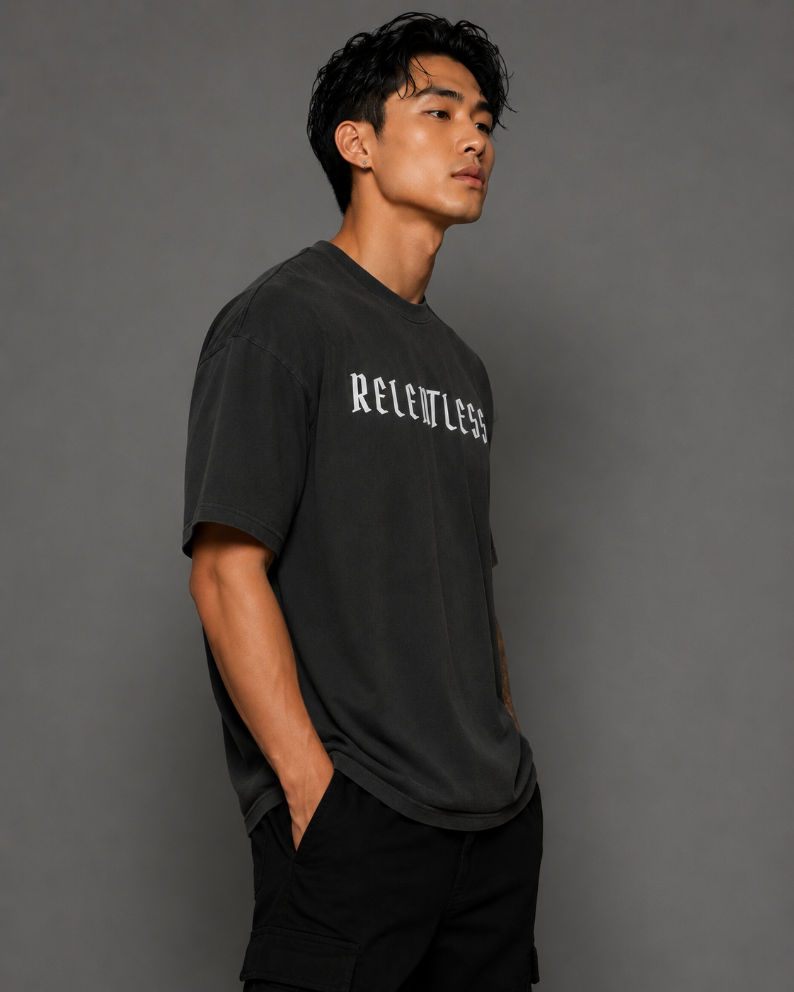 Male model wearing a charcoal oversized “RELENTLESS” graphic T-shirt with black cargo pants against a gray studio background.