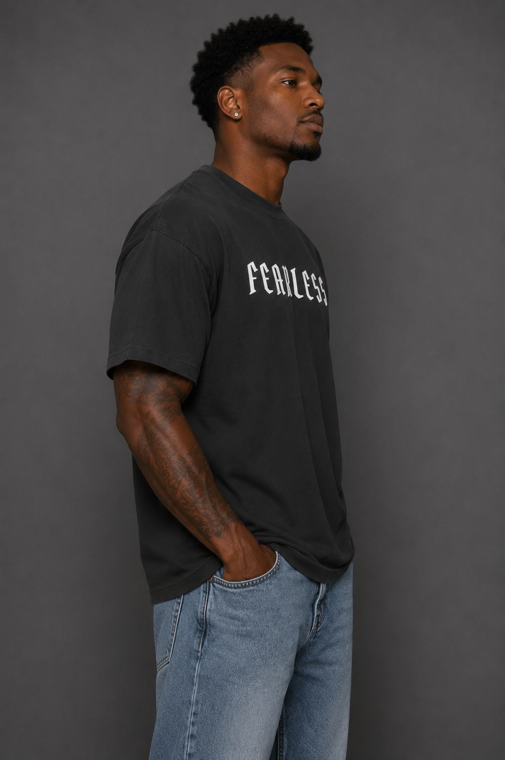Male model wearing a charcoal “FEARLESS” graphic T-shirt and blue jeans in a side-view studio shot against a gray background.