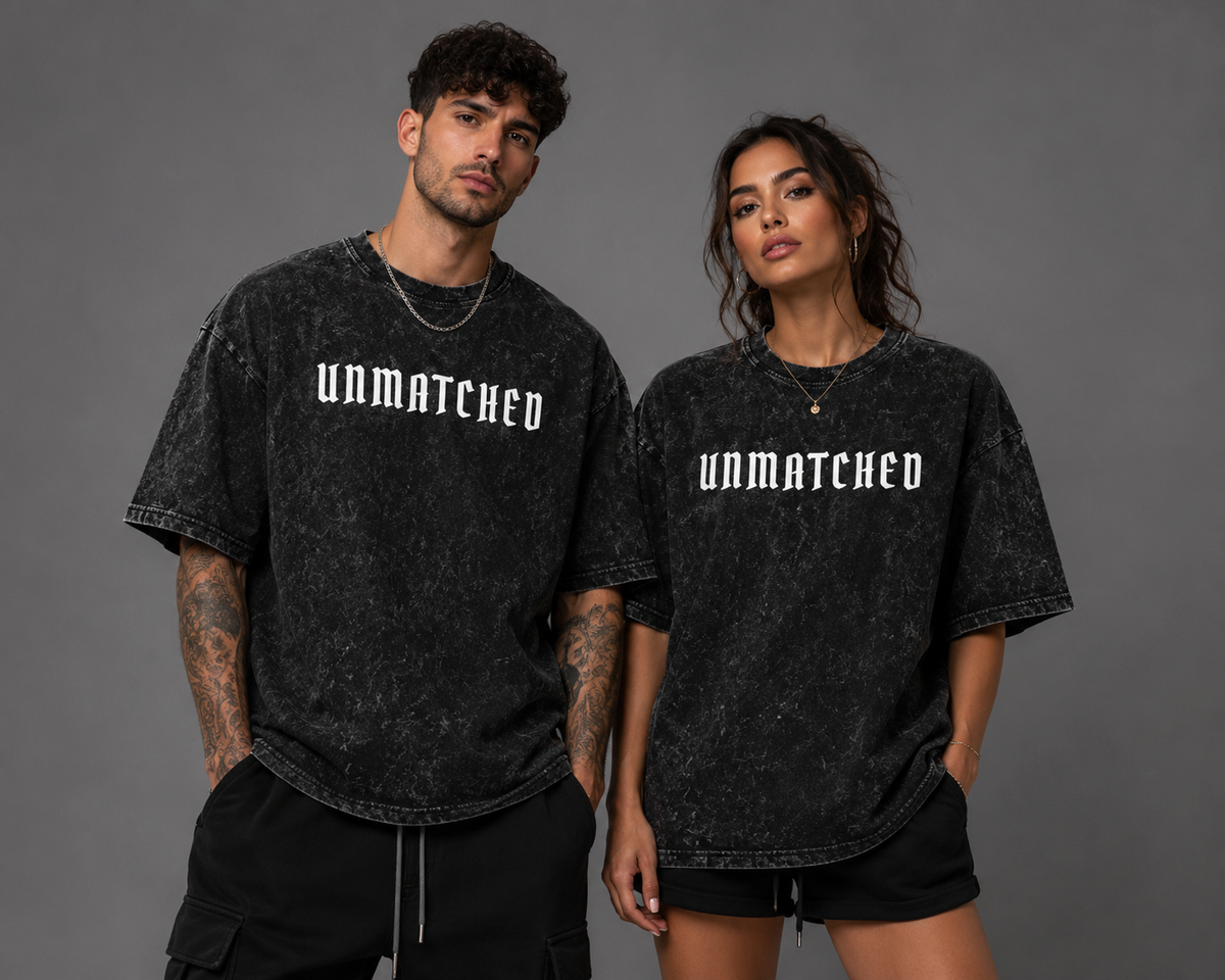 Male and female models wearing black acid-wash “UNMATCHED” graphic T-shirts with black bottoms, posing against a gray studio background.