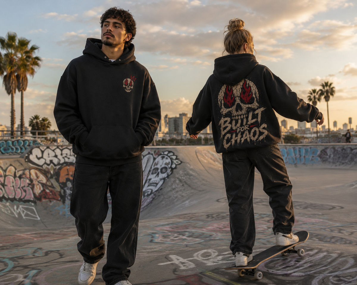 Male and female models wearing black “Built for Chaos” hoodies at a graffiti-covered skatepark, with the female model skateboarding and showing the bold back graphic.