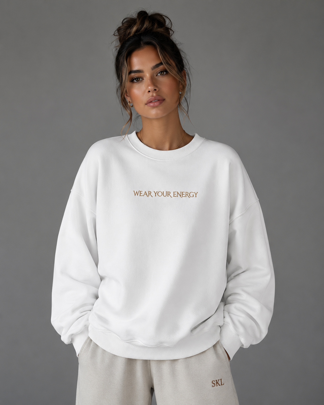 Female model wearing a white oversized “Wear Your Energy” sweatshirt with subtle gold lettering, styled with neutral SKL sweatpants.