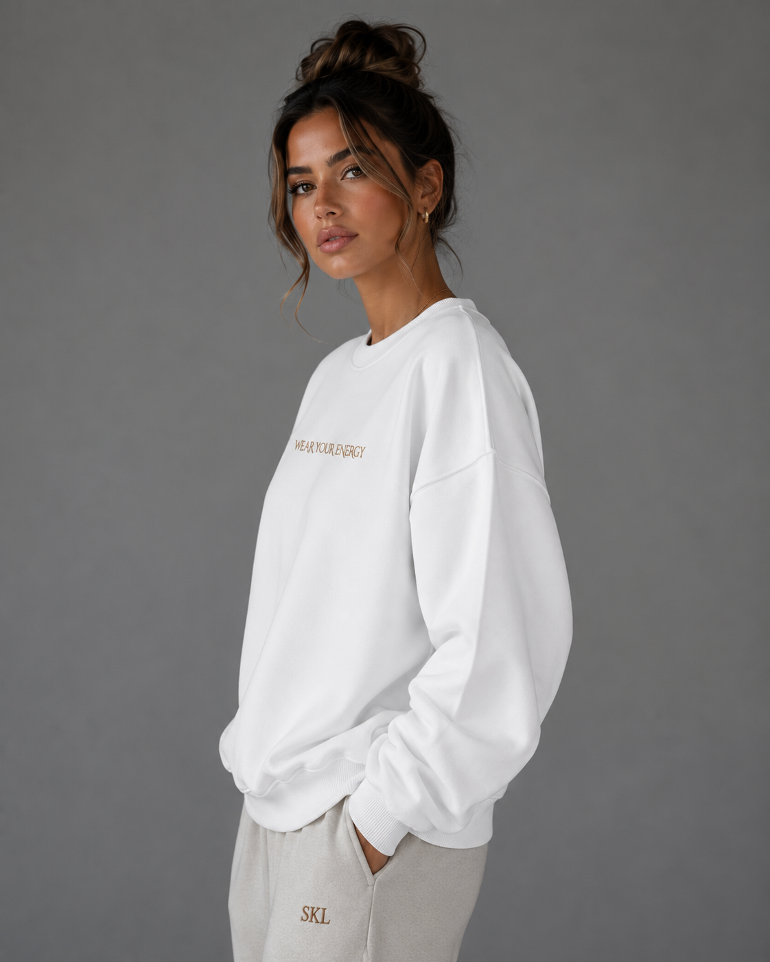 Side view of a female model wearing a white oversized “Wear Your Energy” sweatshirt with subtle gold lettering and neutral SKL sweatpants.