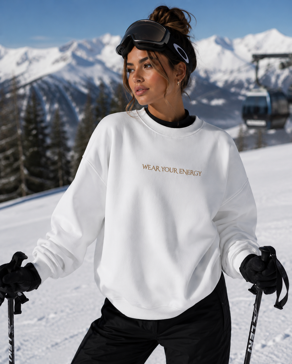 Female model wearing a white oversized “Wear Your Energy” sweatshirt with subtle gold lettering while skiing in the snowy mountains.