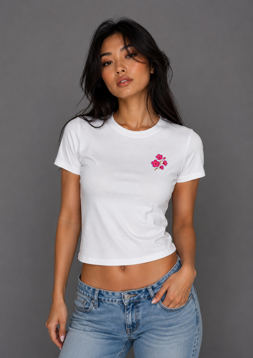 Woman modeling a fitted white cropped T-shirt with a small embroidered pink floral design on the chest, paired with light blue jeans.