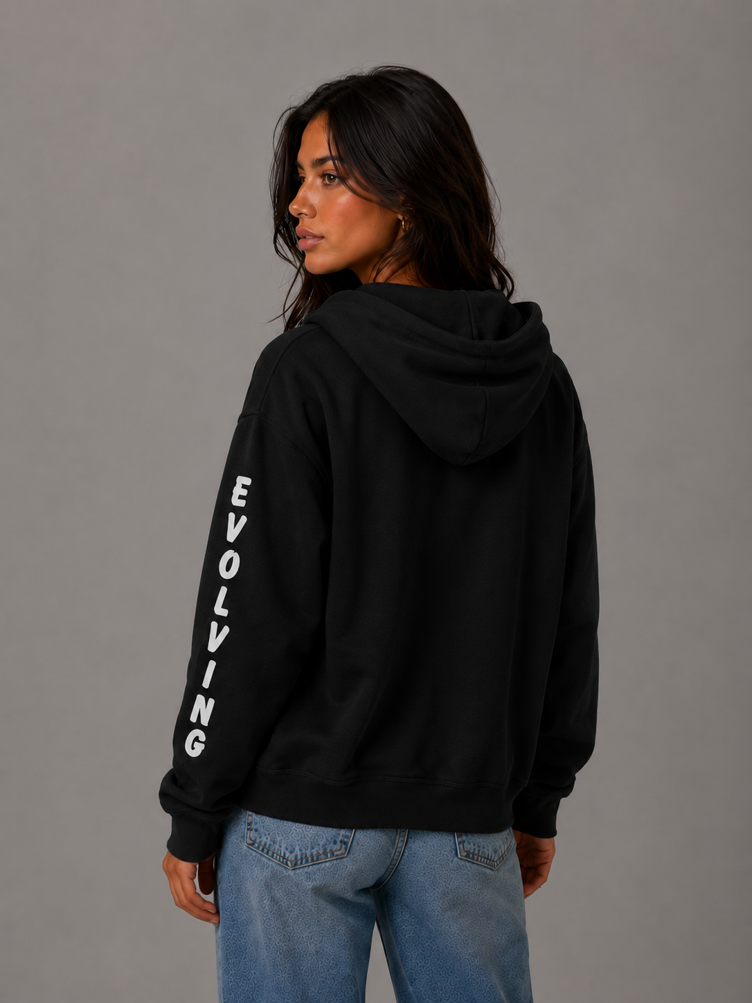Female model showing the back of a black Gently Evolving full-zip hoodie with white “EVOLVING” sleeve lettering, styled with blue jeans.