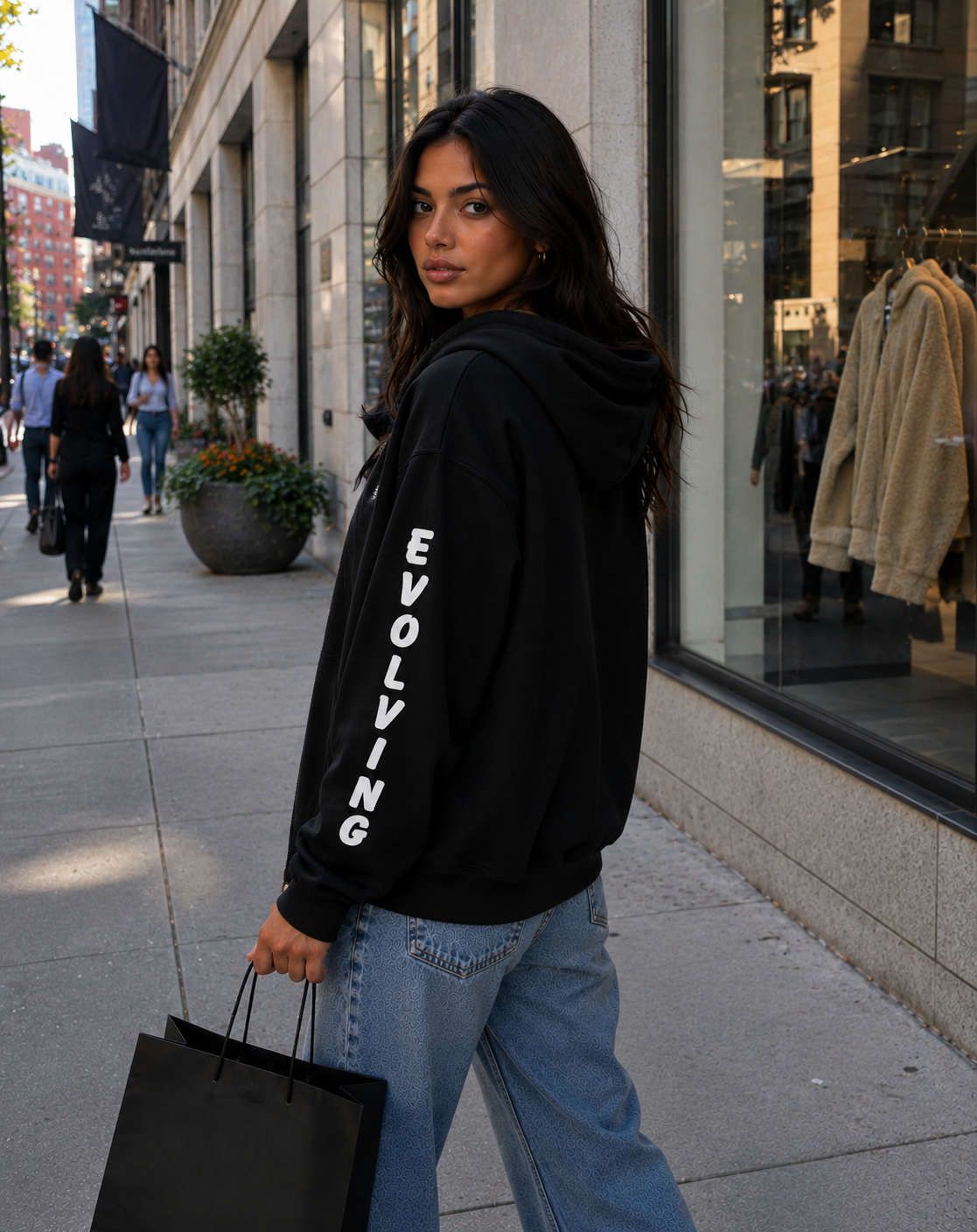 Female model wearing a black Gently Evolving hoodie with white “EVOLVING” sleeve lettering while carrying a shopping bag on a city sidewalk.