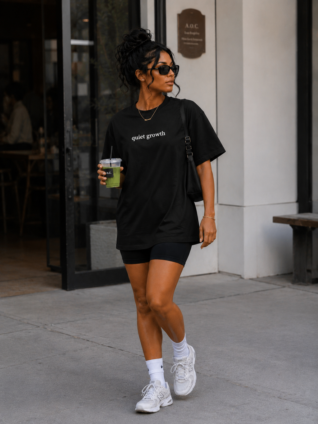 Woman in black t-shirt and shorts holding a drink, walking outside a building.