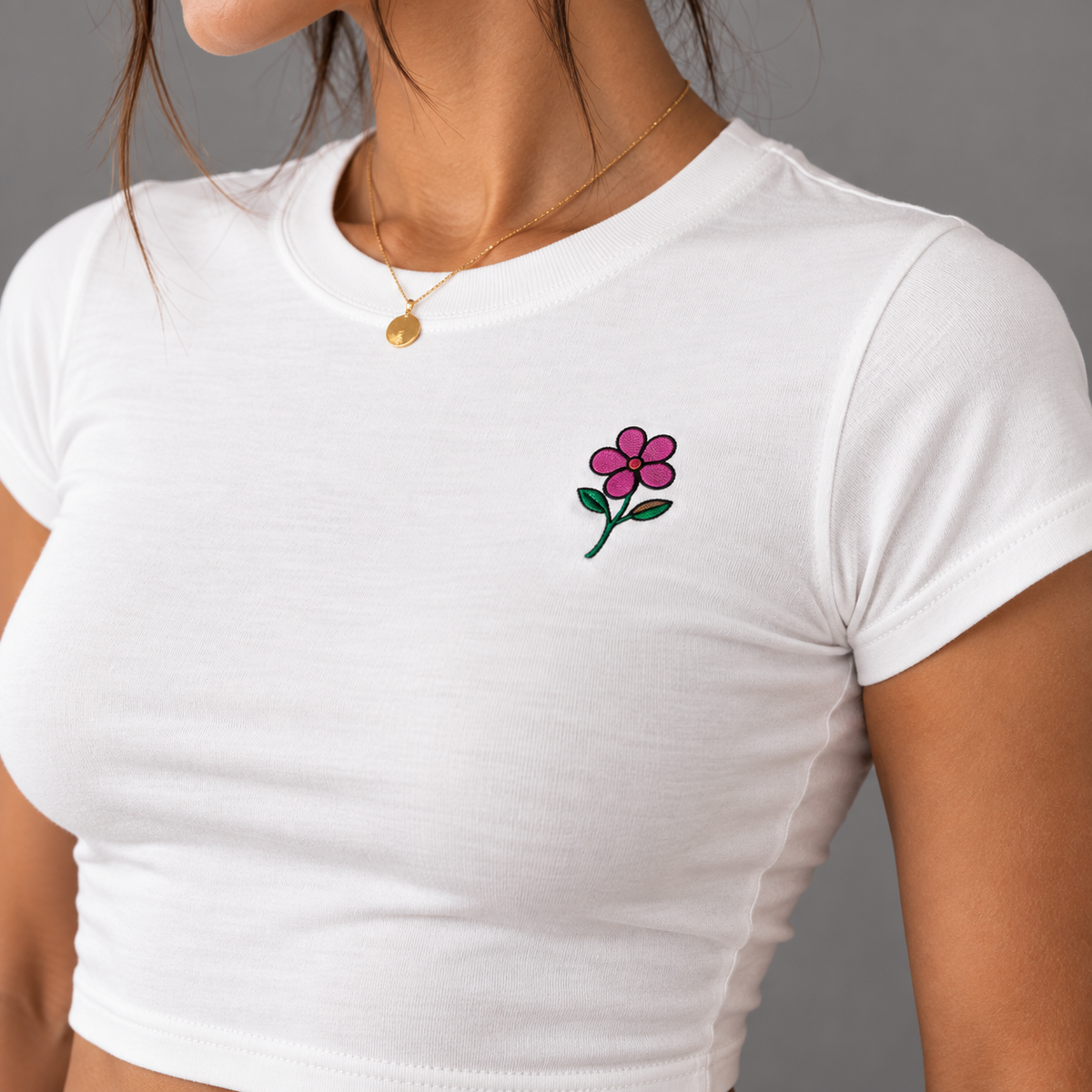 A close-up of a woman wearing a white crop top t-shirt with a small embroidered pink flower on the left chest