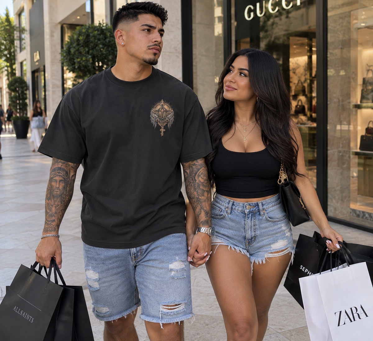 A man and a woman walking hand-in-hand on a shopping mall street, wearing matching black outfits. The man wears a black t-shirt with a gold winged emblem on the chest and distressed denim shorts.