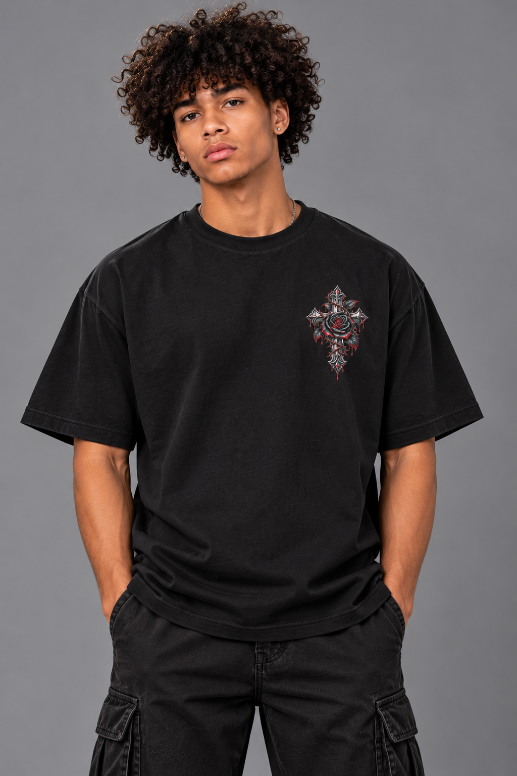 A man with curly dark hair wearing a black short-sleeve t-shirt with a colorful embroidered cross graphic on the left chest, paired with black cargo pants, against a solid gray studio background.