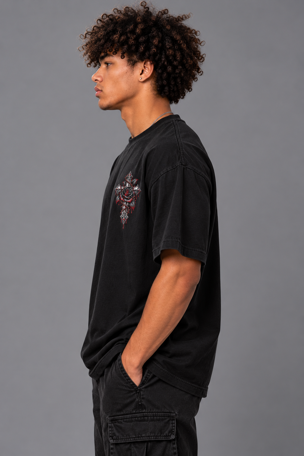 A man with curly dark hair is shown in profile wearing a faded black t-shirt with a red and white graphic on the left chest, paired with black cargo pants, against a solid gray studio background.