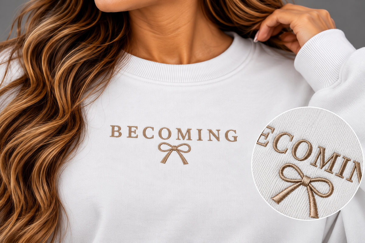 A close-up of a woman wearing a white crewneck sweatshirt with 'BECOMING' and a bow graphic embroidered in tan on the chest. A circular inset highlights the embroidery detail.