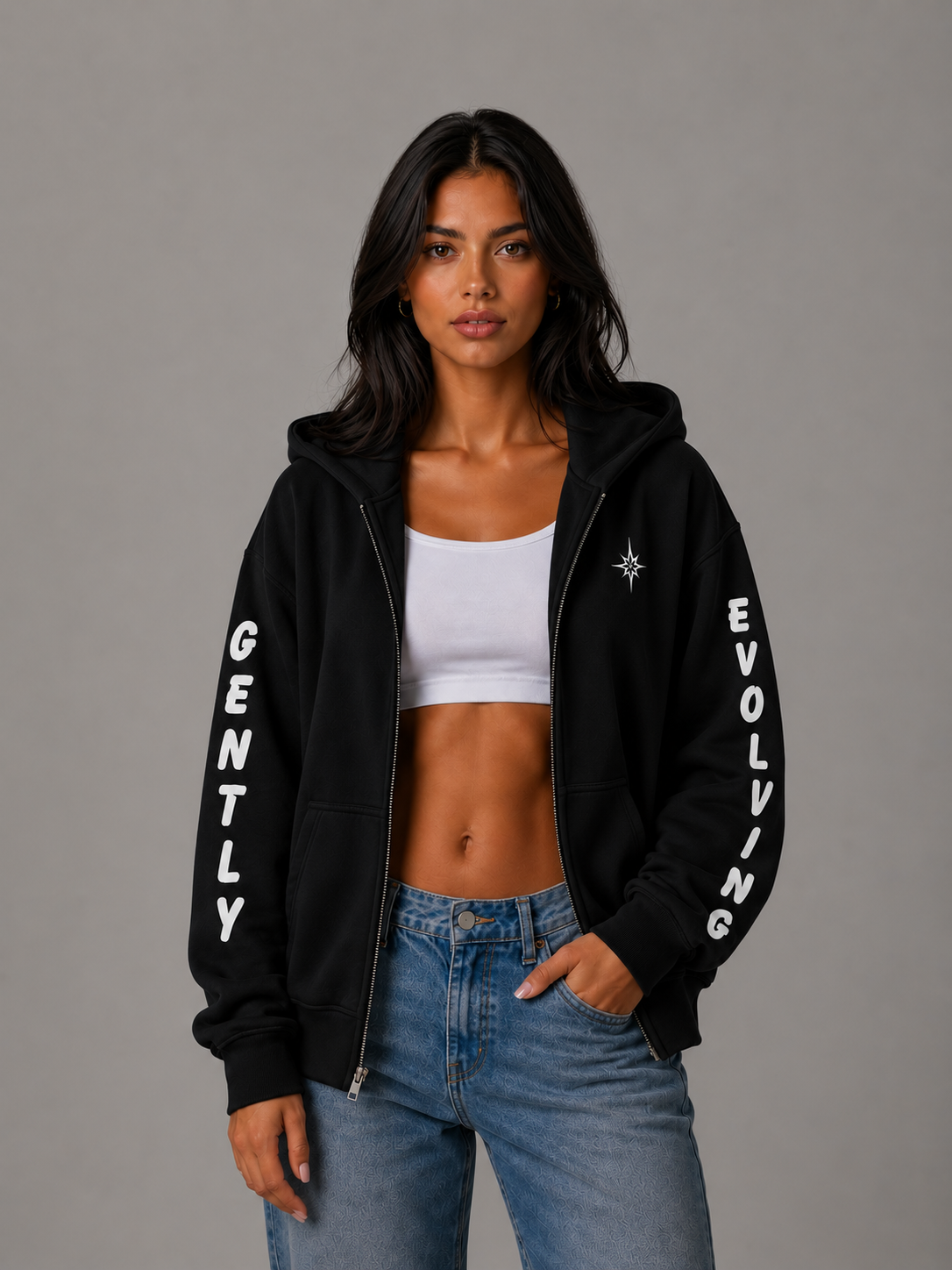 Female model wearing a black Gently Evolving full-zip hoodie with white sleeve lettering and embroidered star detail, styled with blue jeans.