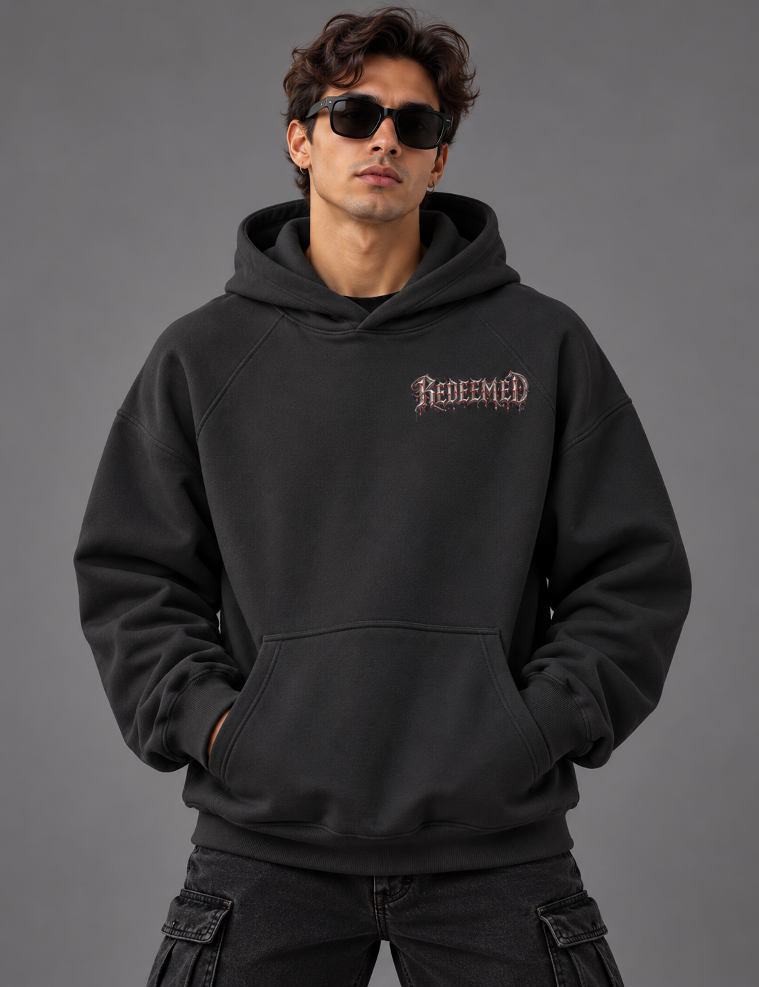 A man wearing a black pullover hoodie with a gothic-style 'Redeemed' logo on the chest, styled with black cargo pants and sunglasses against a solid gray studio background.
