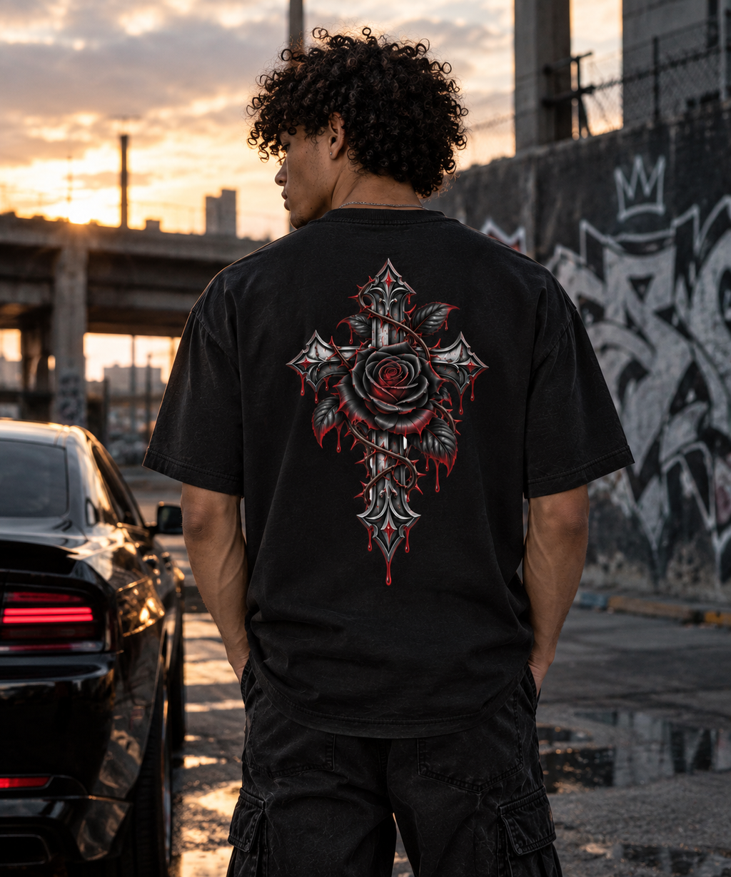 A man viewed from behind wearing a black t-shirt with a large gothic cross and rose graphic on the back, standing in an urban street setting at sunset.