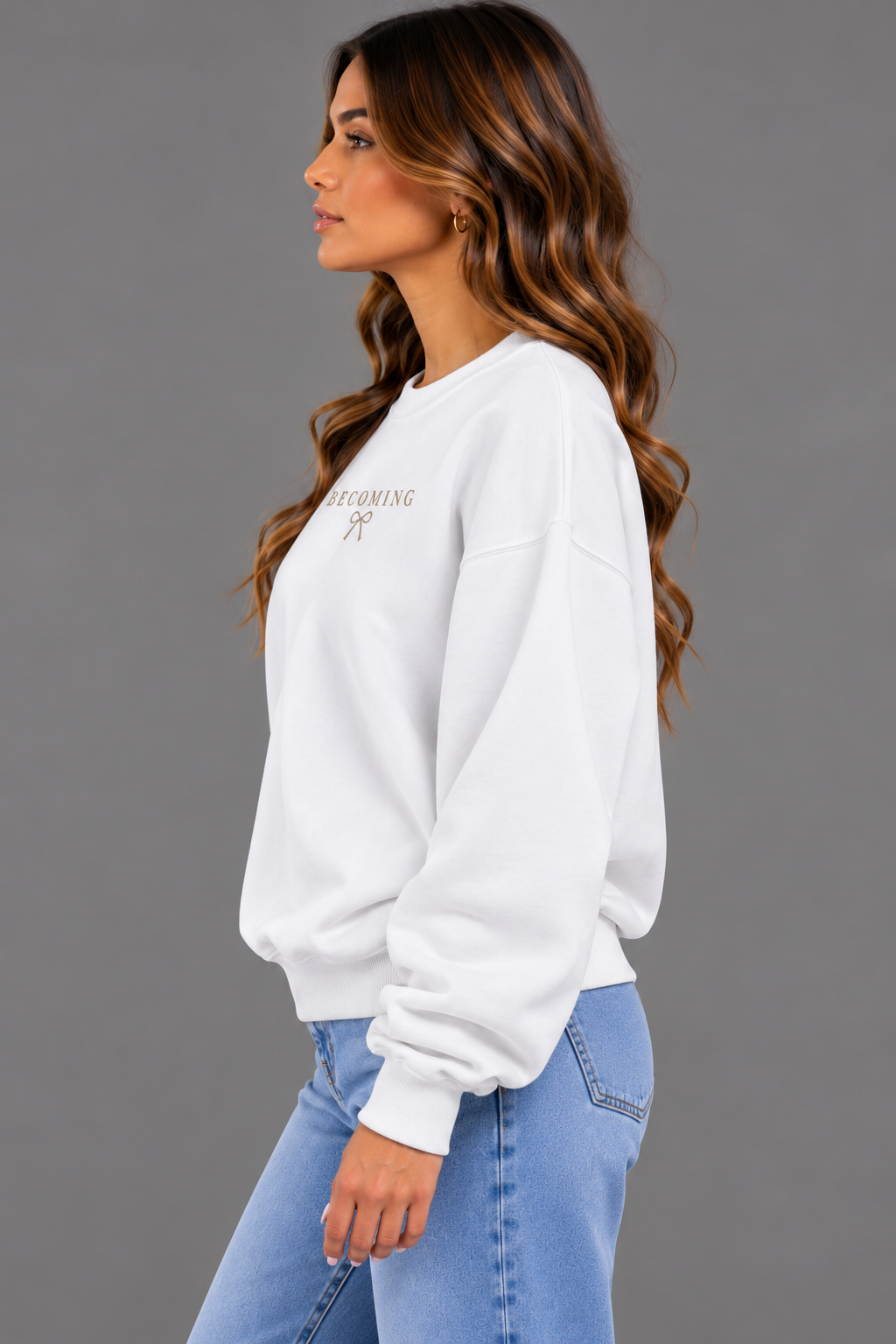 A woman in a white crewneck sweatshirt with gold text reading 'BECOMING 23' and light blue jeans, shown in profile against a solid gray background.