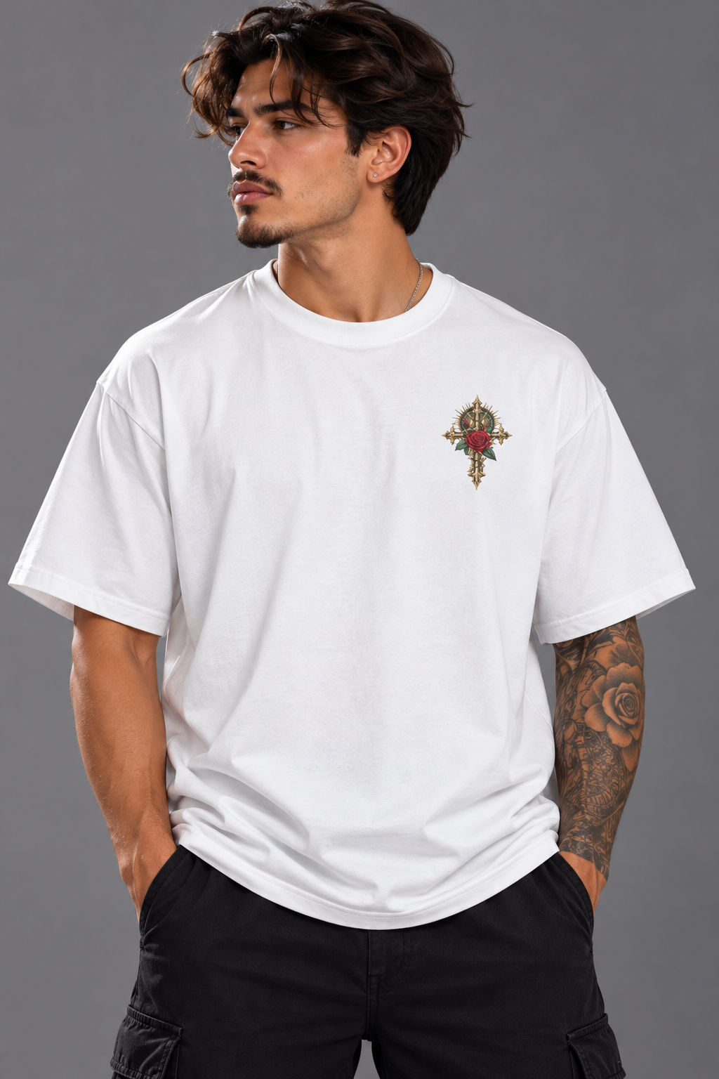 A man wearing a white crewneck t-shirt with a small ornate cross graphic on the left chest, paired with black cargo pants, against a solid gray studio background.