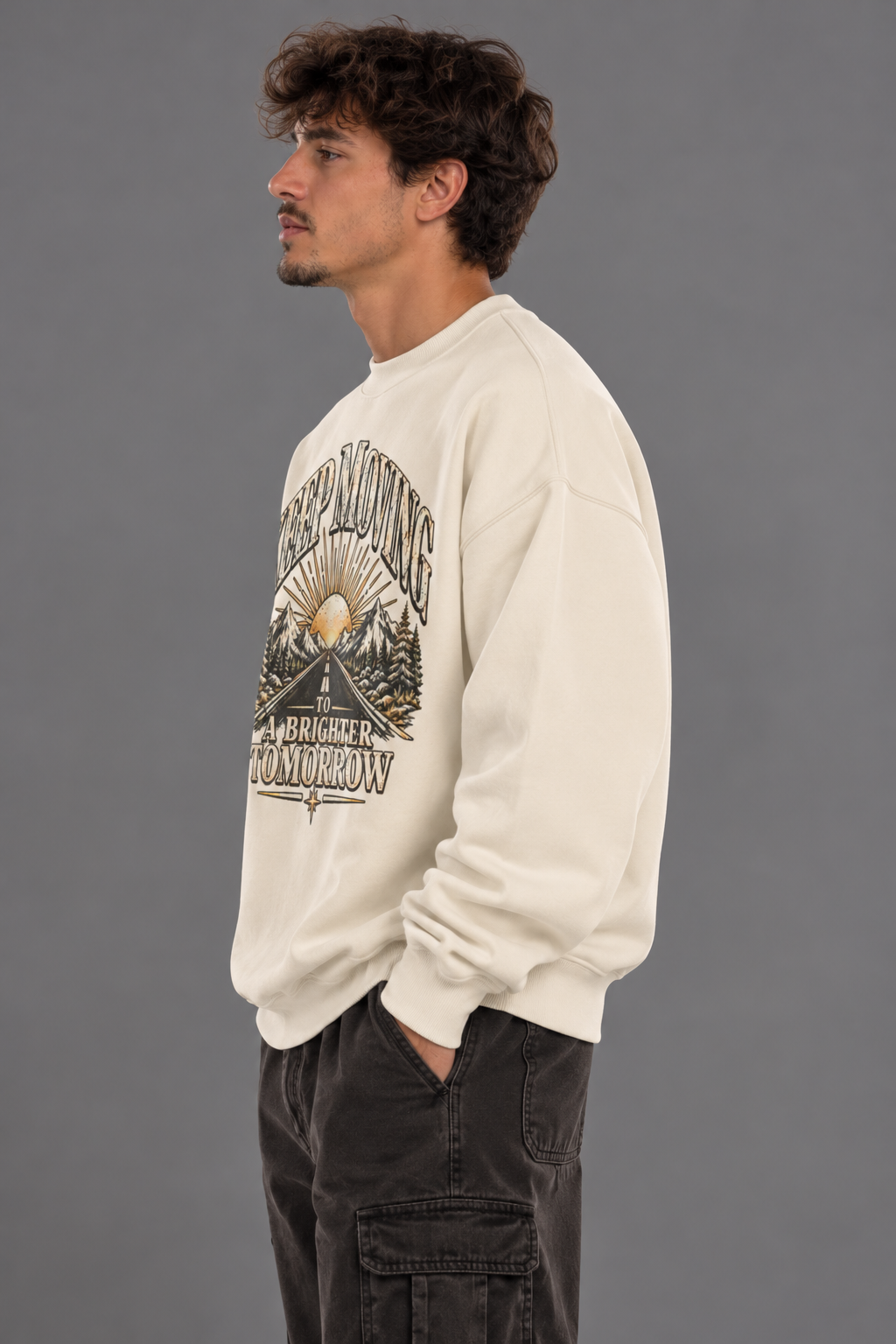 A man wearing a cream-colored crewneck sweatshirt with a detailed black and gold mountain graphic on the front, paired with dark cargo pants, standing against a solid gray studio background.
