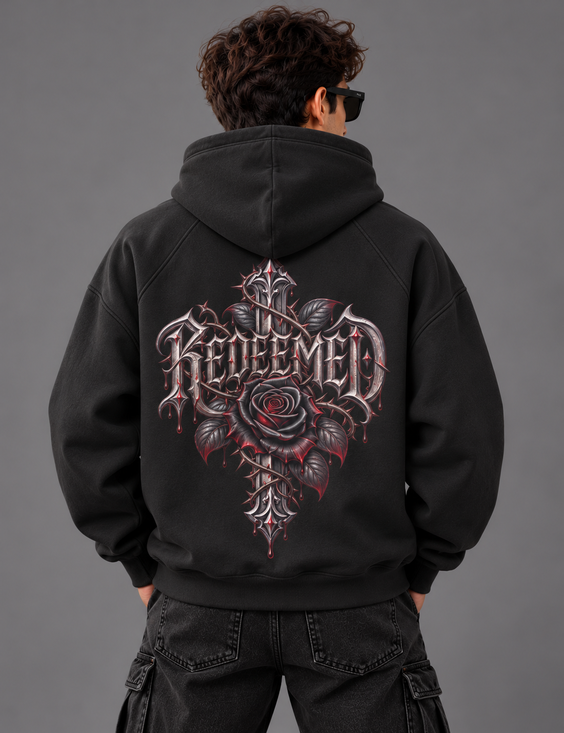 A person shown from the back wearing a black hoodie with a large gothic-style graphic on the back featuring a rose and the word 'Redeemed' in ornate lettering, against a plain gray studio background.