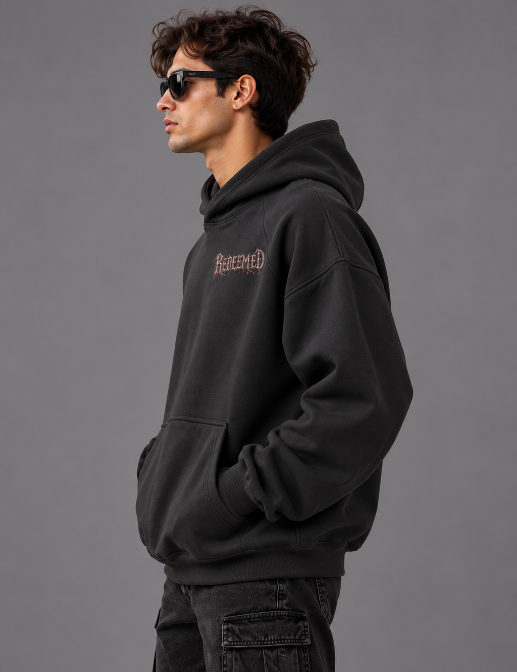 A man wearing a faded black pullover hoodie with a distressed pink 'Reclaimed' logo on the chest, paired with dark gray cargo pants and sunglasses, photographed against a solid gray studio background.