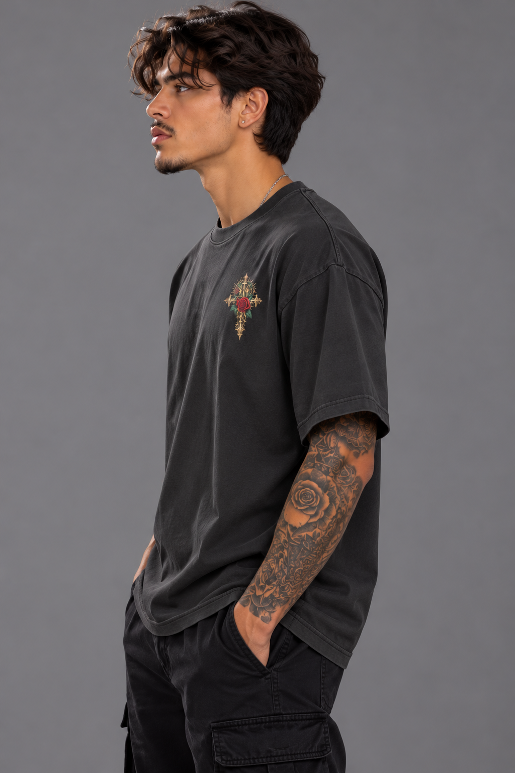 A man wearing a faded black crewneck t-shirt with a small colorful cross graphic on the left chest, paired with black cargo pants, photographed against a solid gray studio background.