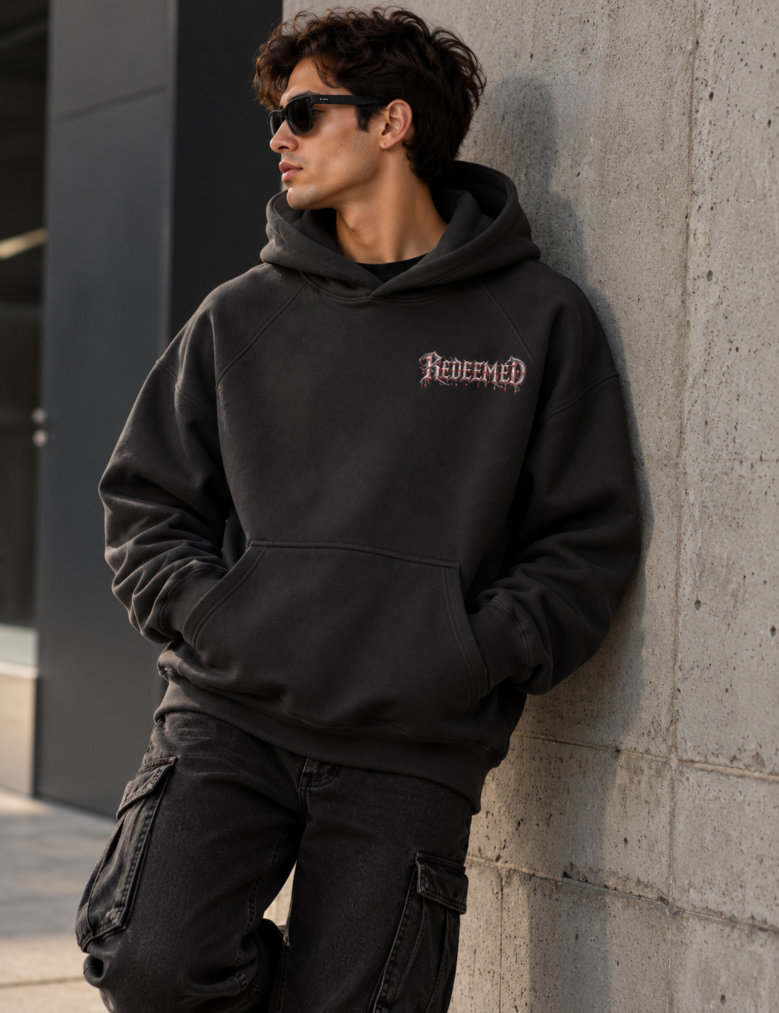 A man wearing a faded black pullover hoodie with 'Redeemed' embroidered in a gothic font on the chest, leaning against a concrete wall.